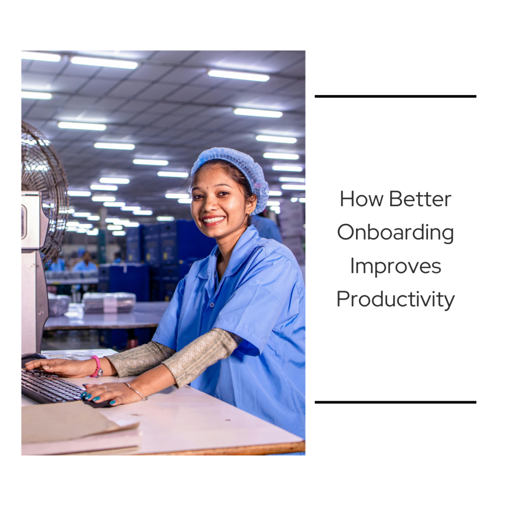 A smiling female worker in a blue lab coat and hair net uses a computer workstation on a light industrial manufacturing floor.