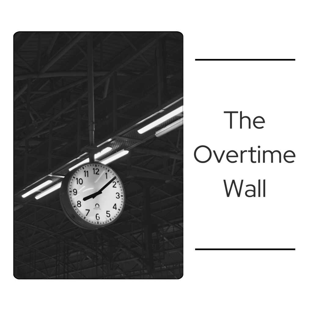 Black and white photo of a large clock hanging from the steel ceiling beams of an industrial building, a reminder of the hours behind mandatory overtime.